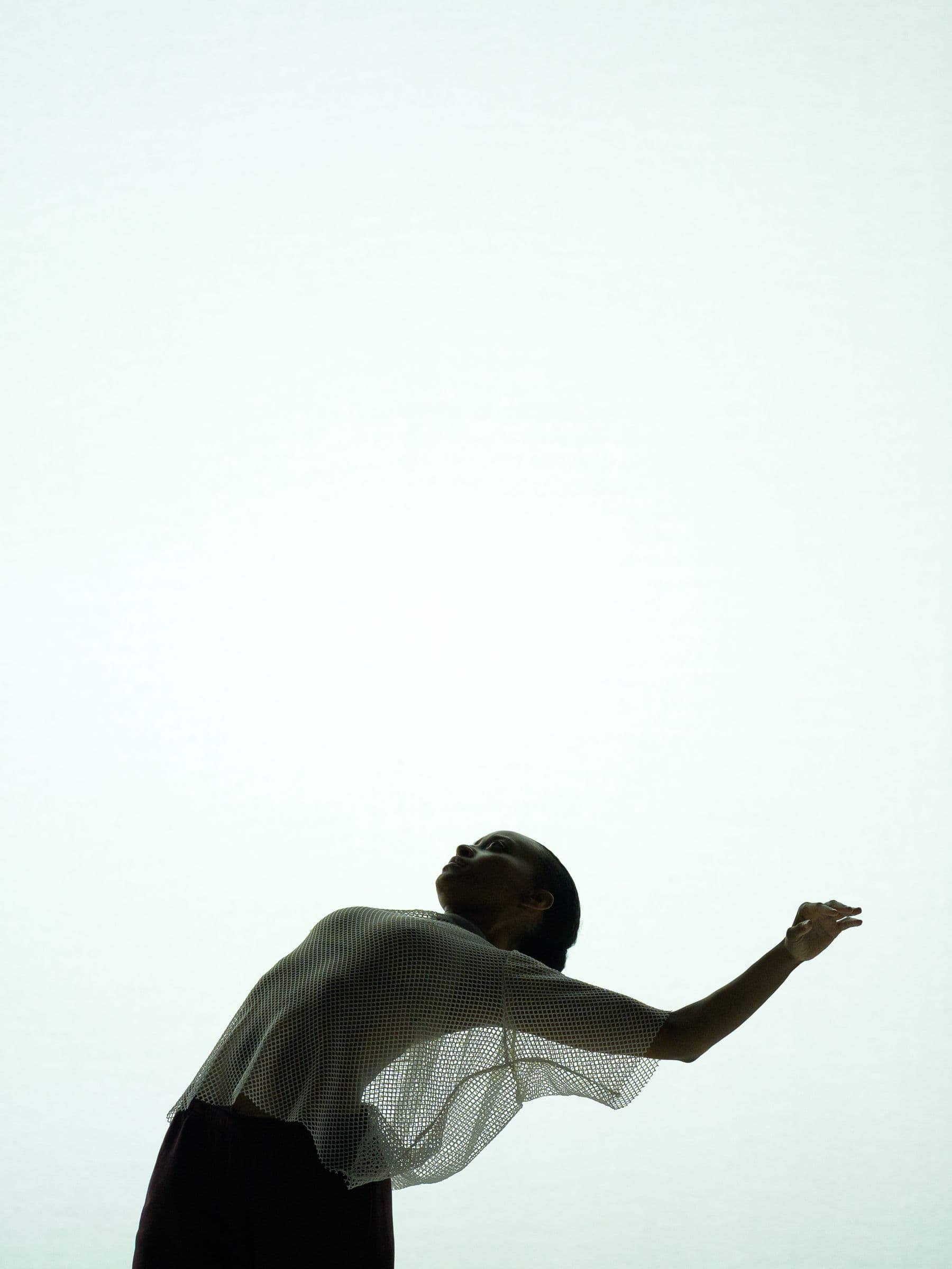 Faith Joy Mondesire silhouetted in a mesh top leans back with head tilted up and arm outstretched, set against a pale, minimalist backdrop