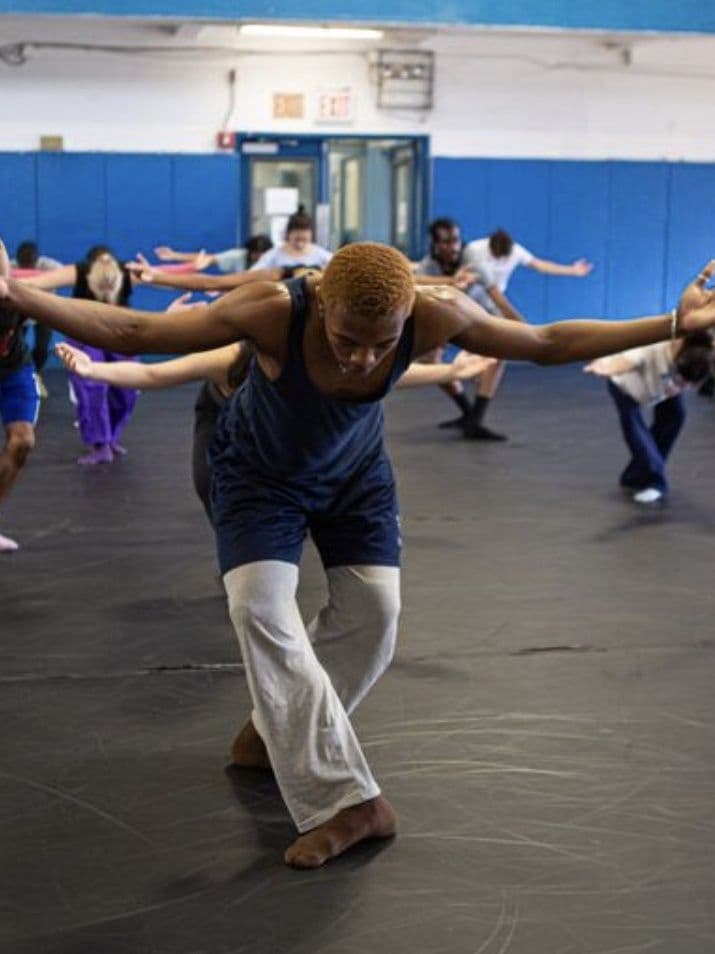 A group dance class in a studio with blue padded walls, dancers reaching their arms out in unison as an instructor in front leads the movement, leaning forward with arms spread wide.